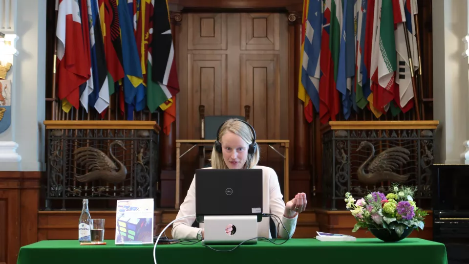 Sofie in front of her laptop in front of the flags in the IIIEE aula. Photo. 