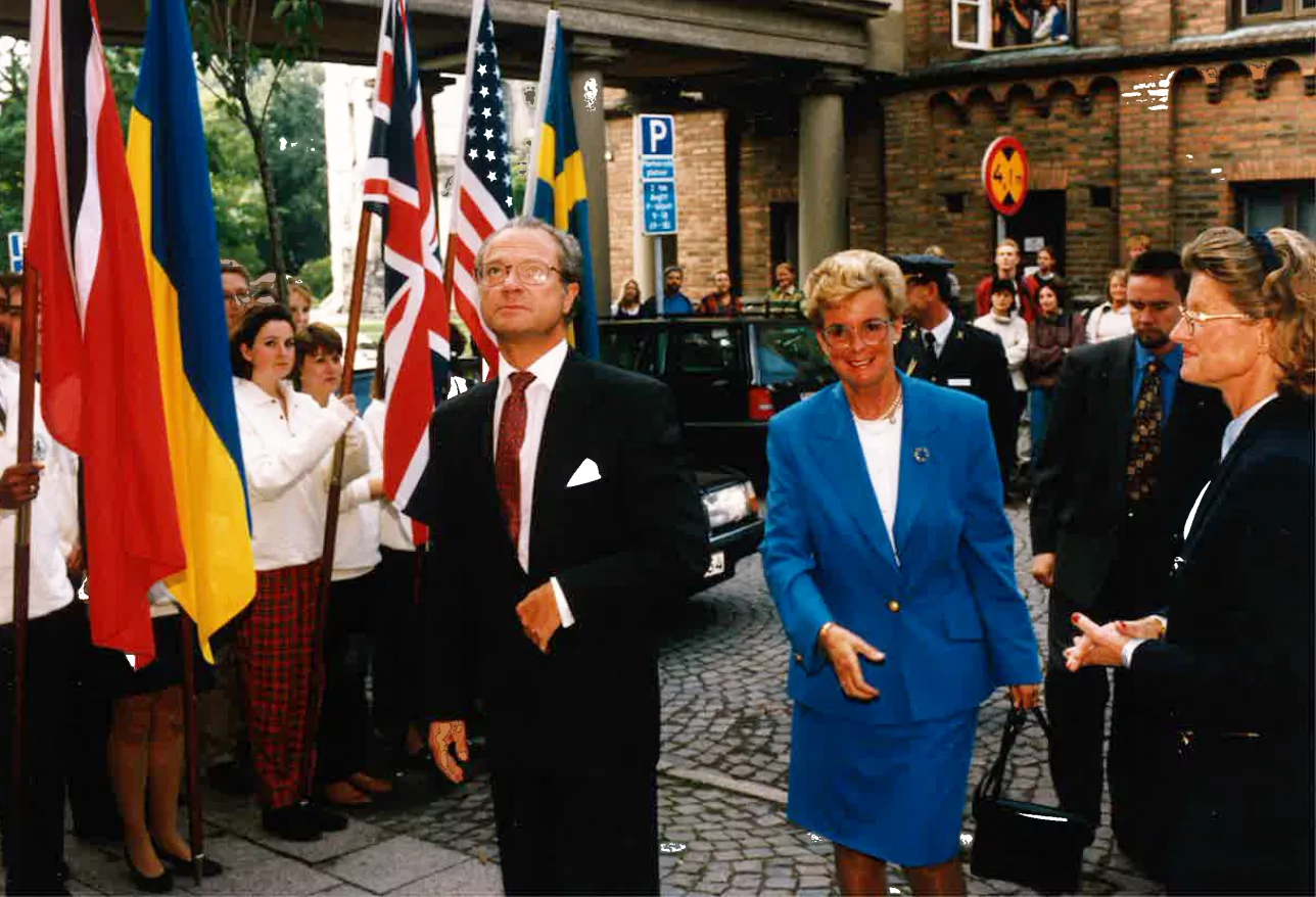 King Carl XVI Gustaf in suit on the street outside the IIIEE. A woman in blue suit next to him and people standing with flags on the sides. Photo. 