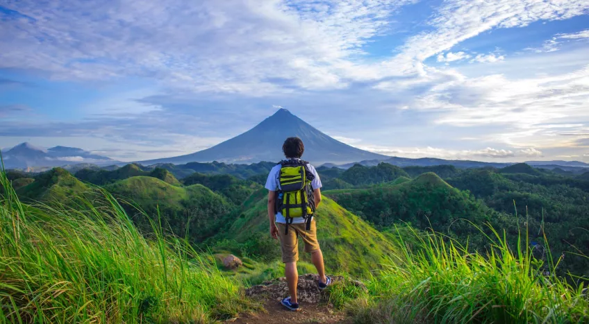 A man wearing shorts and a backpack stands with his back towards the camera. In front of him a mountain rises up in the distance